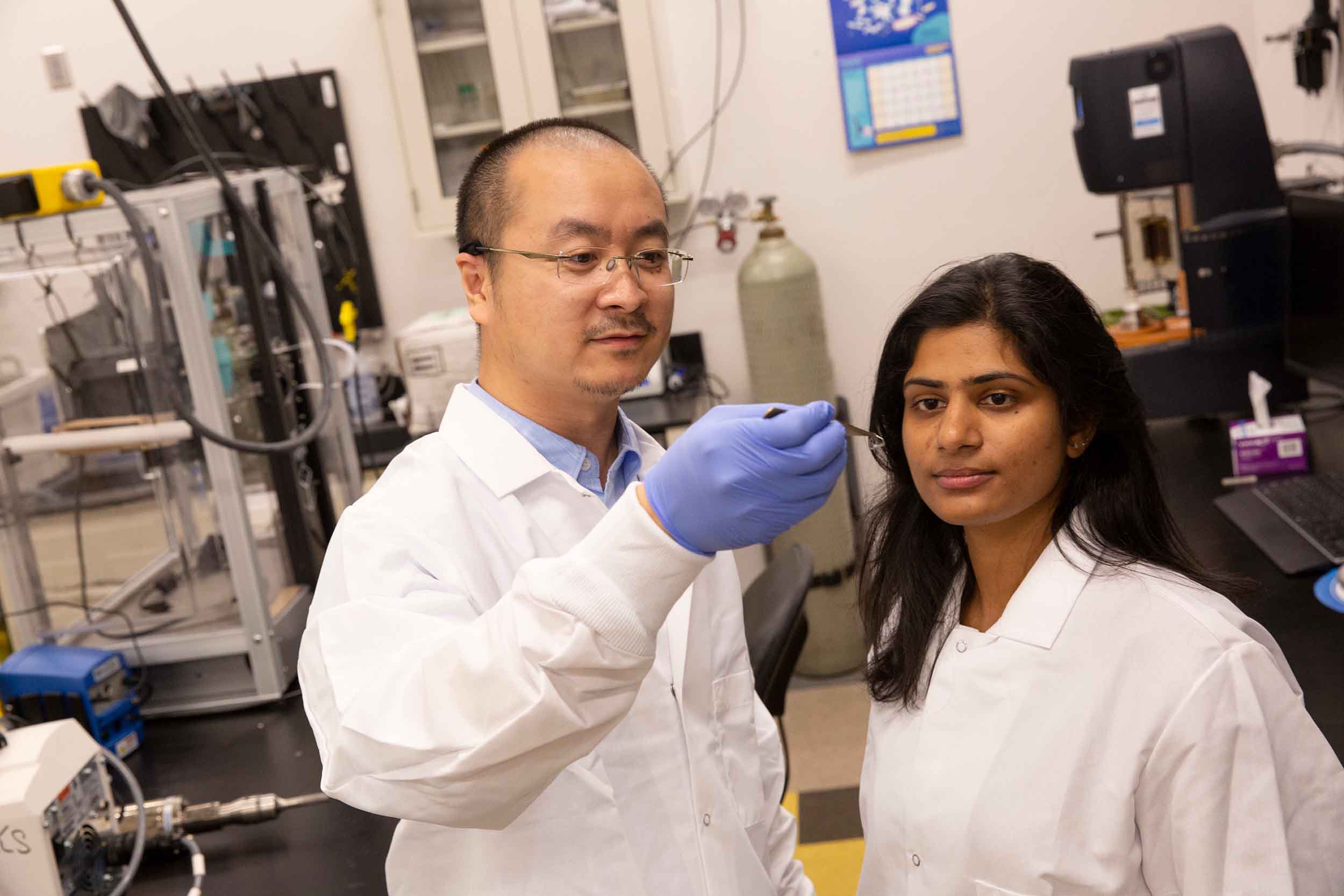 Kenan Song holds a very small 3D printed sample with a pair of tweezers, showing it to his research collaborator, Mounika Kakarla, while in his lab