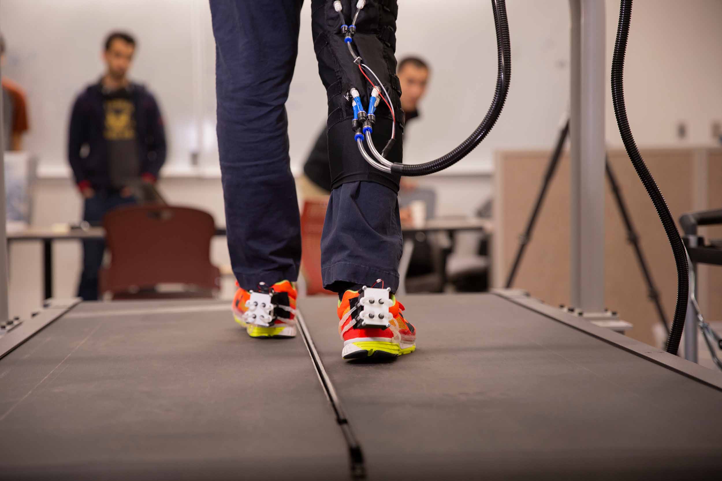 a person wearing robotic devices on their knee and both shoes walks on a treadmill while being observed by researchers