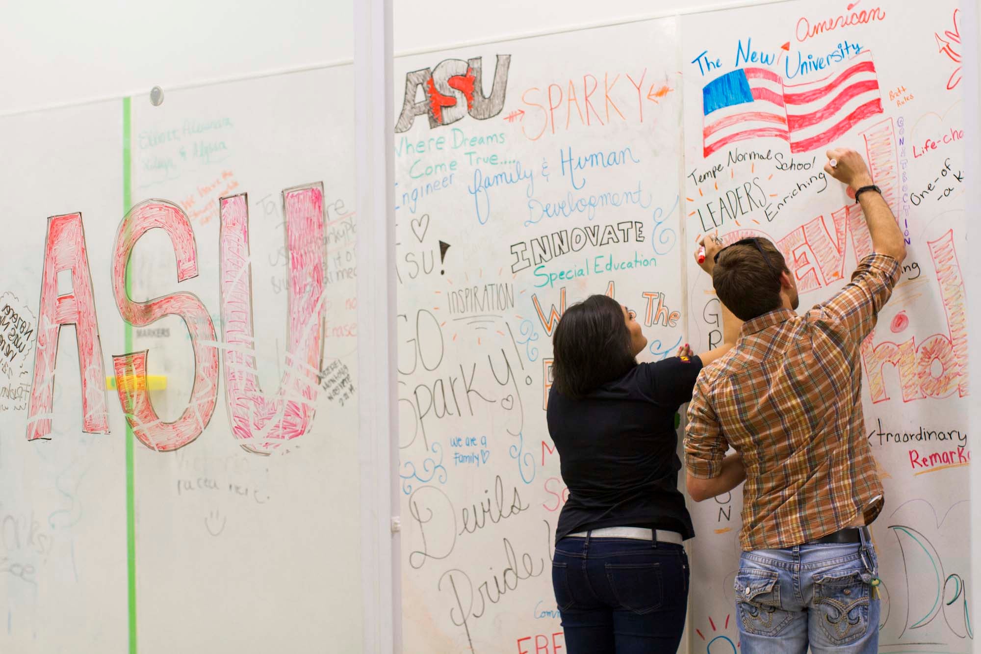A man and woman write on a huge white board wall, where many others have written positive ASU sentiments before