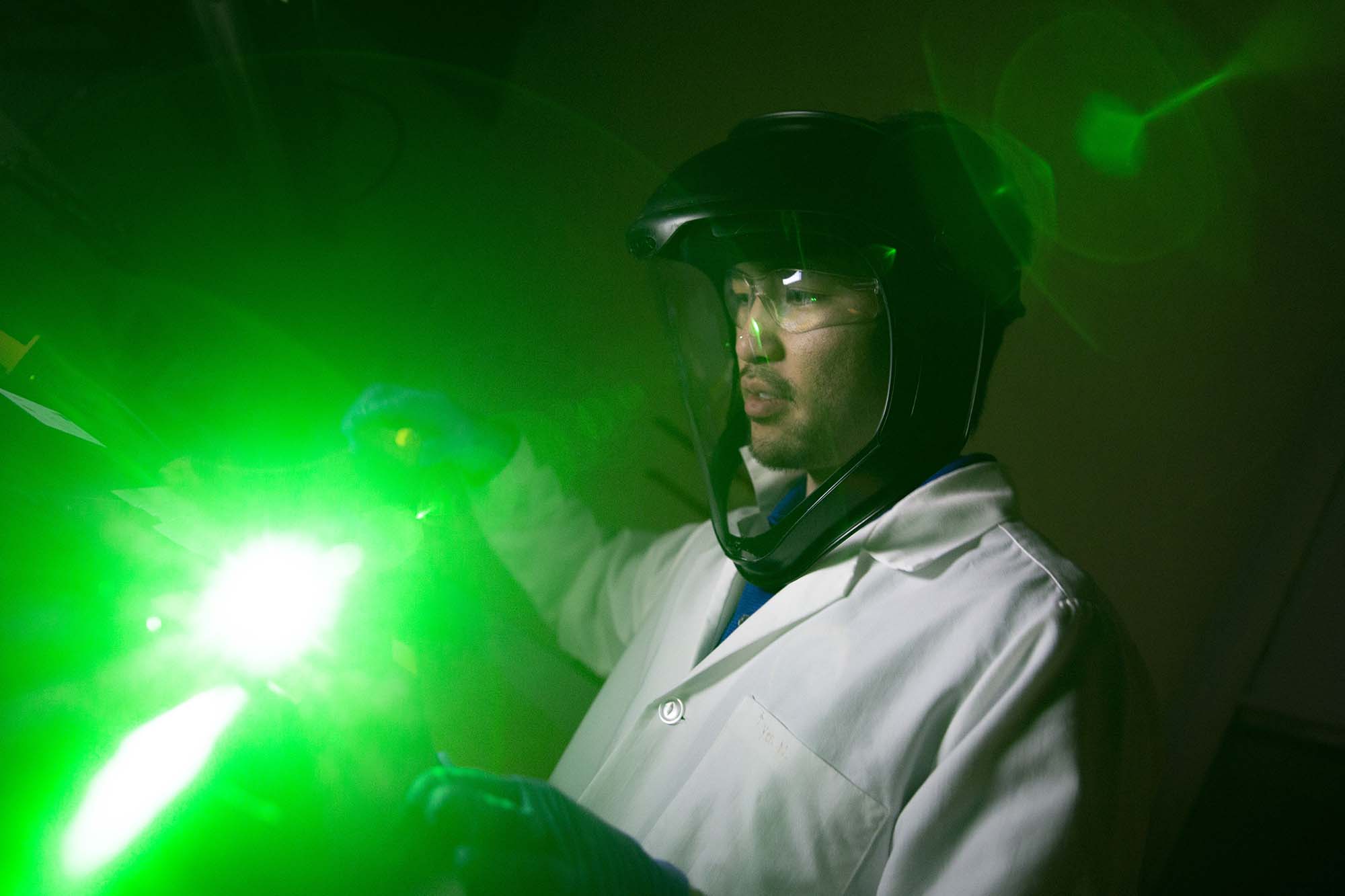 An ASU Engineering student, wearing a lab coat, measures bright green light being emitted in a dark room by germicidal optical fibers.