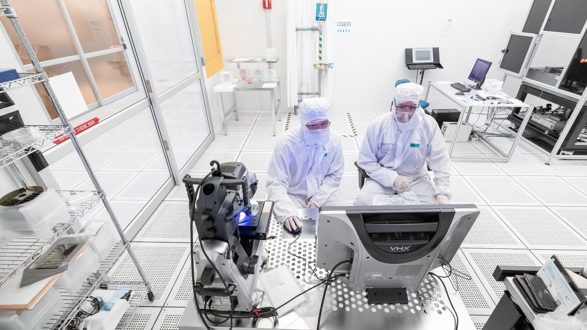 Two people in white lab coats and other protective gear looking at a computer in a lab environment.