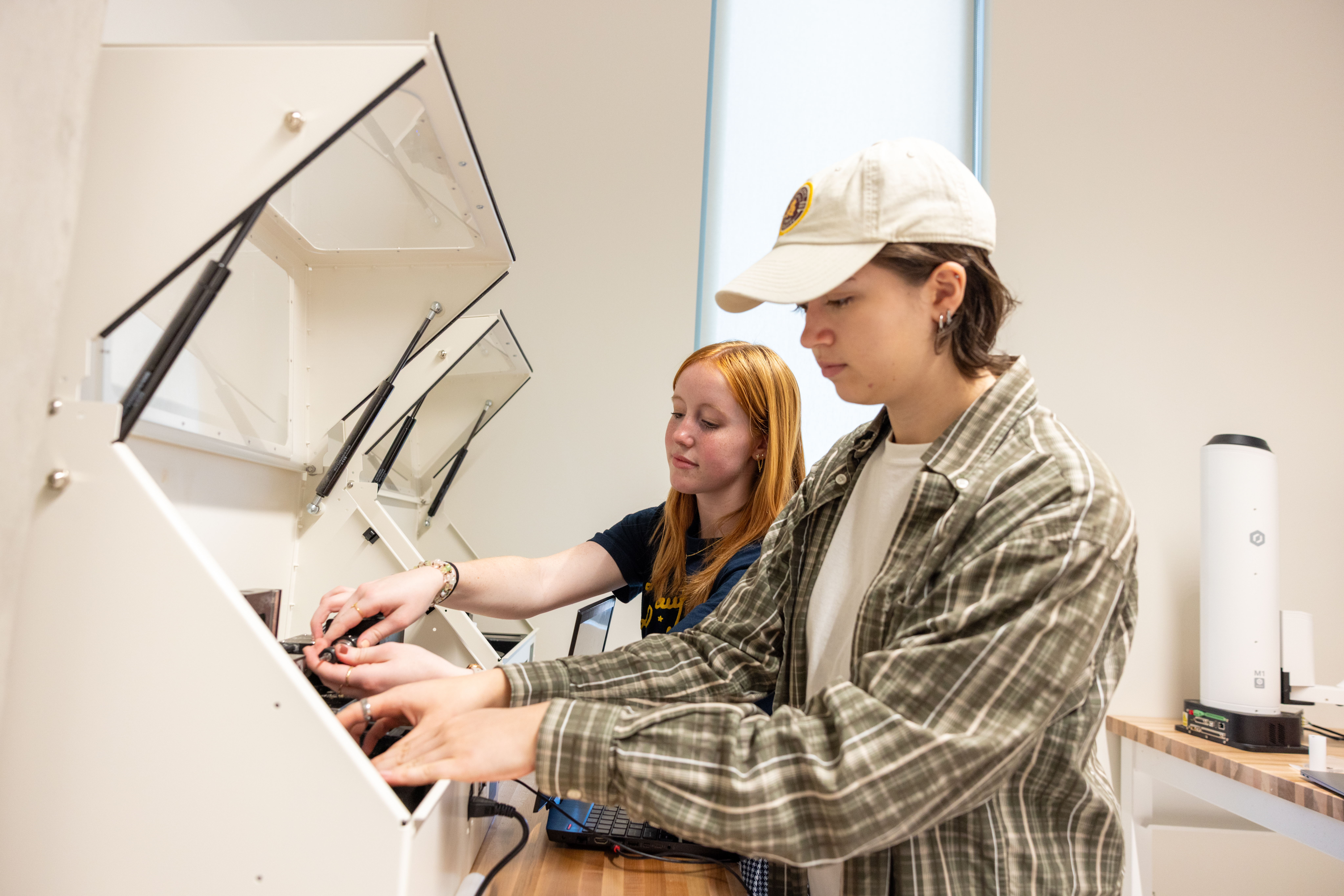 Two students work together at a lab workstation, adjusting components inside an open machine while a laptop sits nearby.