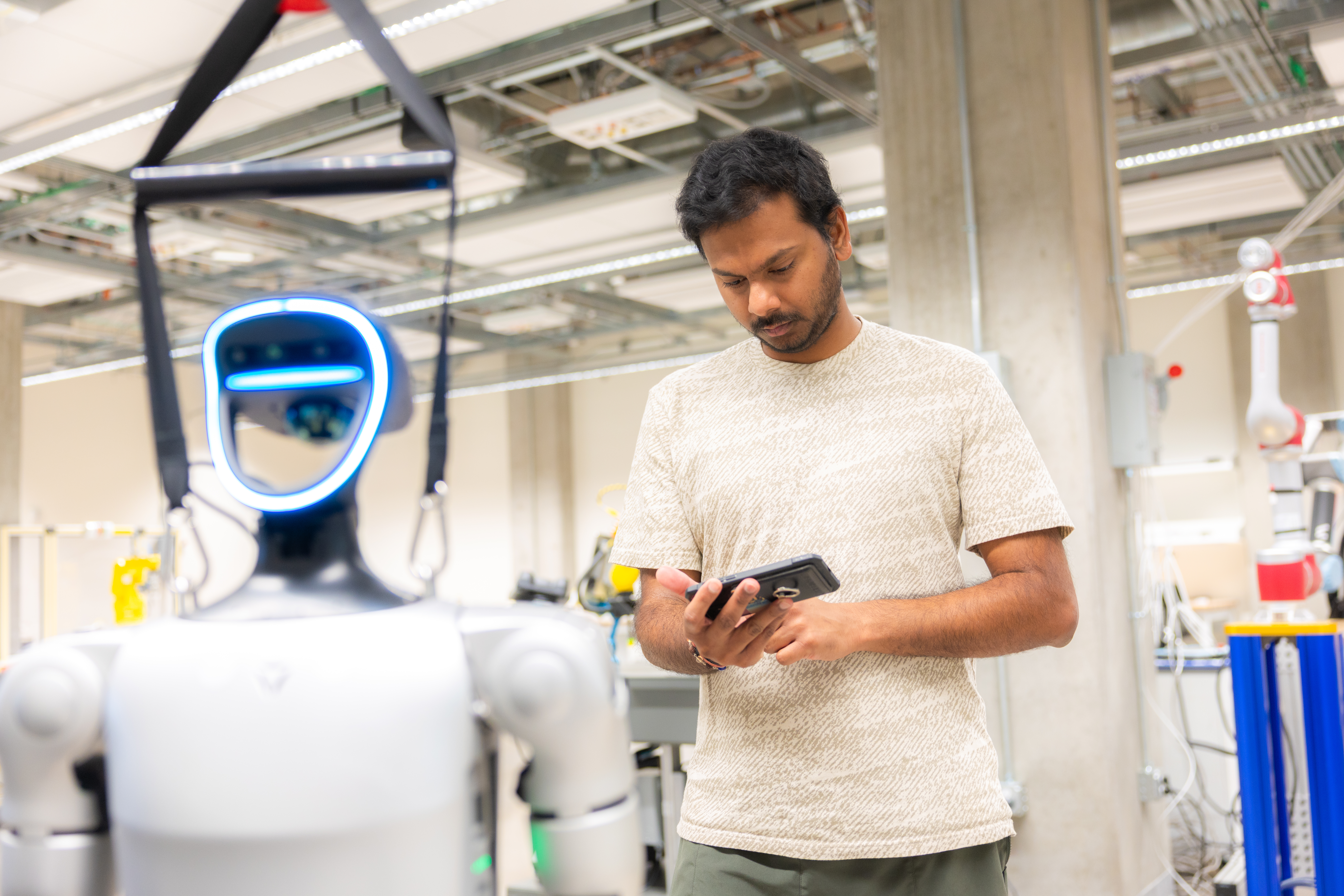 A student stands in a lab using a tablet to control or monitor a humanoid robot positioned nearby.