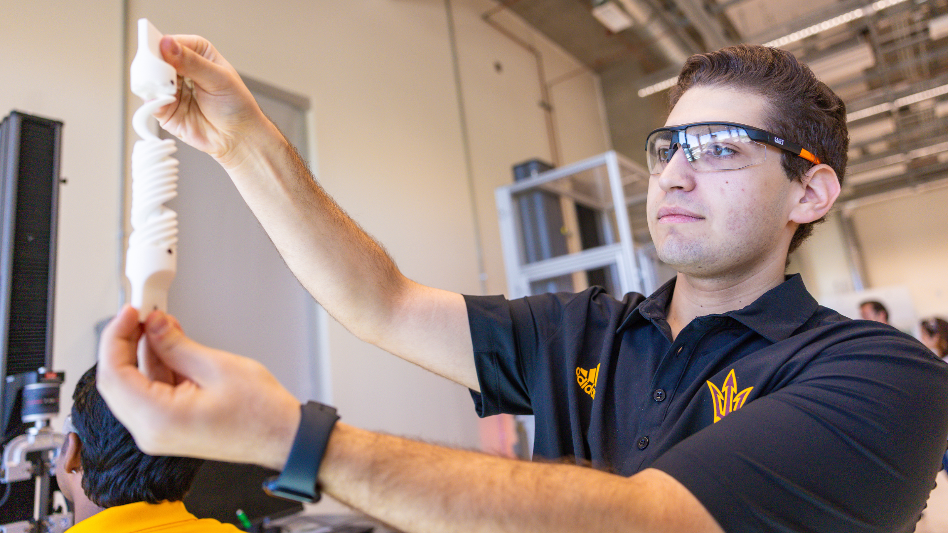 A student wearing safety glasses in a lab holds up a white, flexible, spiral-shaped 3D-printed object, examining it closely.
